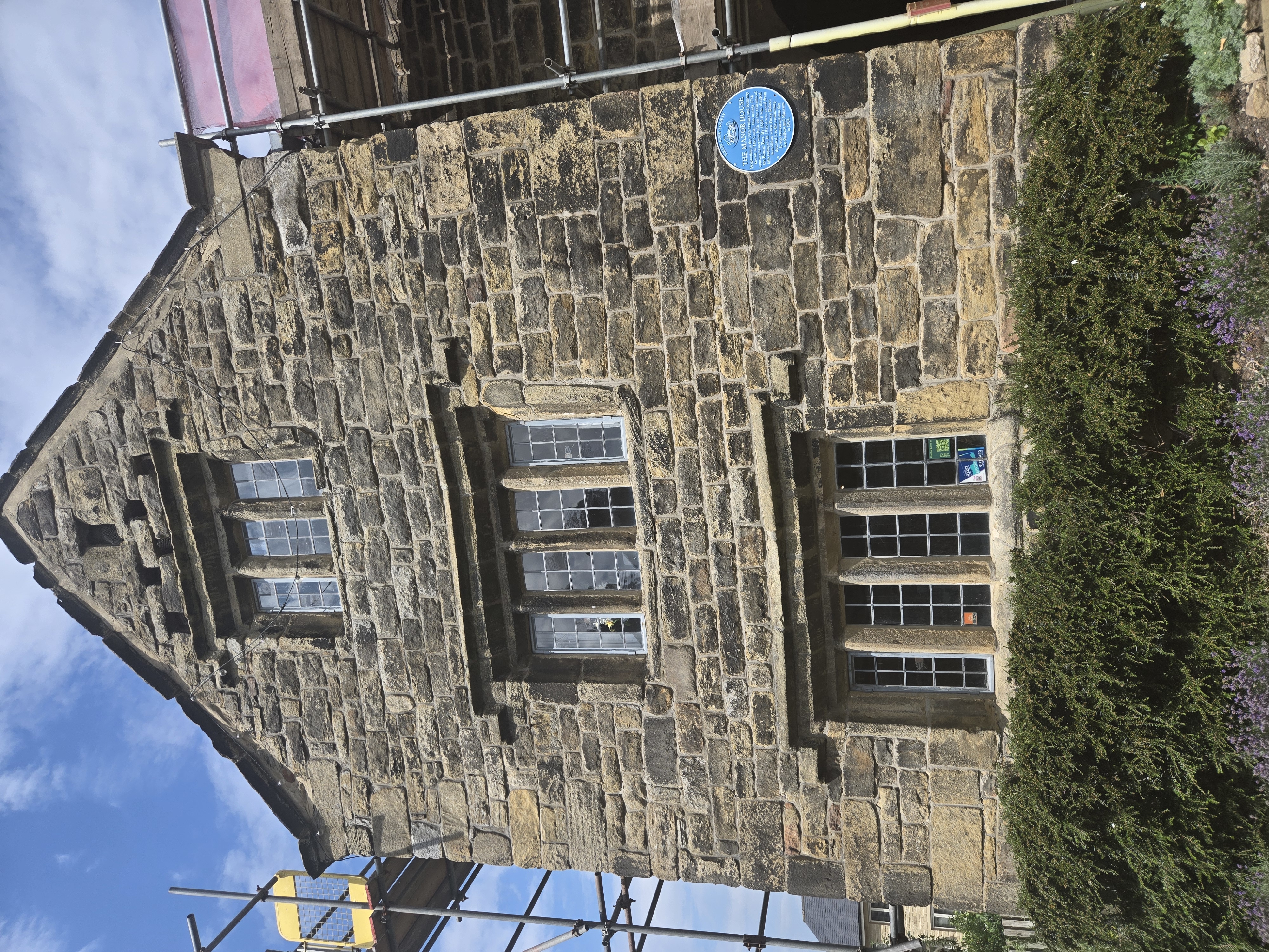 Front of dark stone faced medieval building with blue plaque adjacent to door and mullioned windows